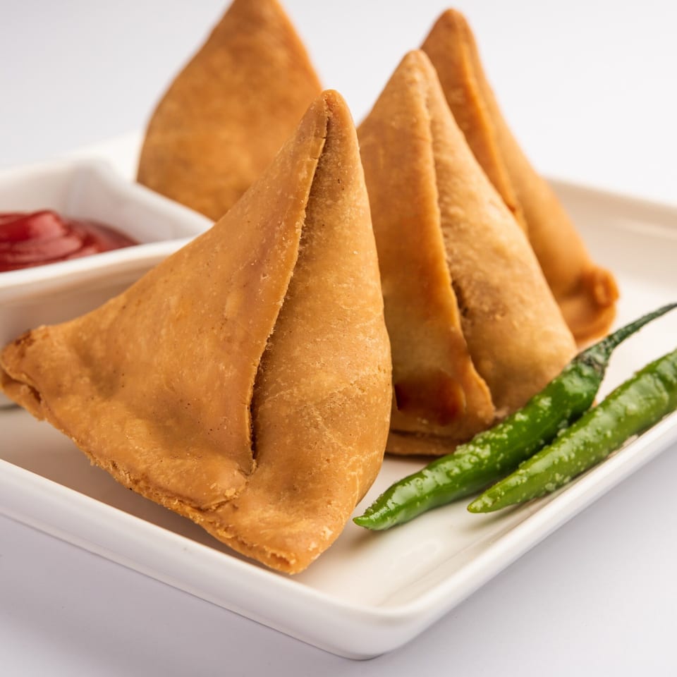Golden-brown samosas, a popular Indian appetizer, served with ketchup and green chilies on a white plate.