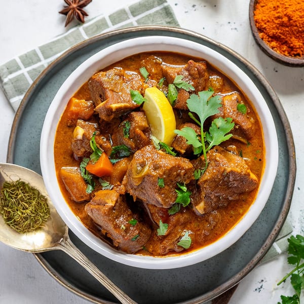 Indian lamb curry with lemon, cilantro, and spices in a white bowl on a gray plate.