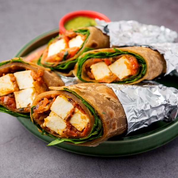 Indian paneer kathi rolls, some wrapped in foil, on a green plate with a side of green chutney.