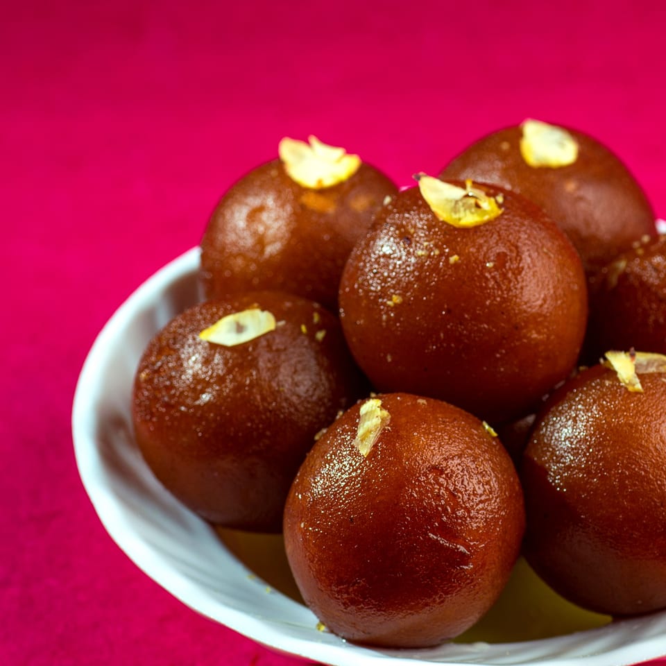 A white bowl filled with several glistening gulab jamun, garnished with slivered almonds, against a pink background.