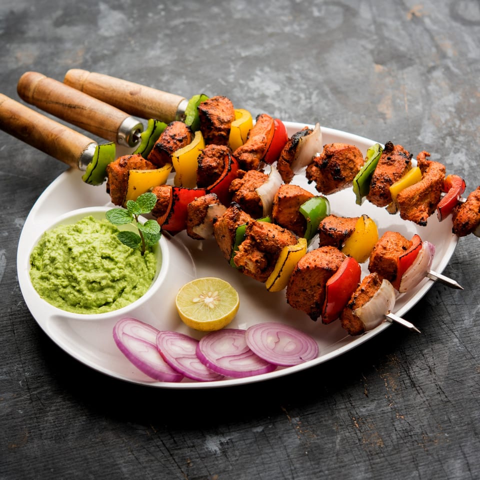 Three skewers of Indian chicken tikka with bell peppers and onions, served with green chutney, lemon, and red onion.