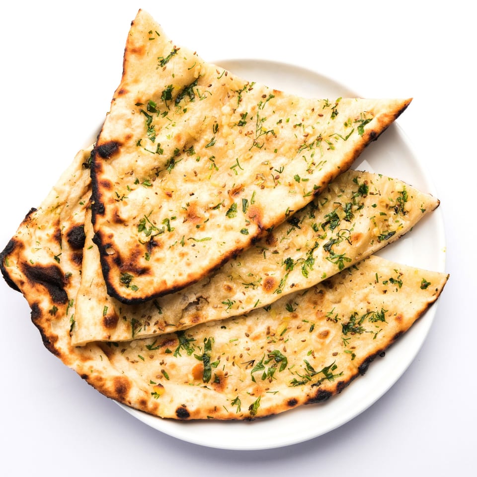 Golden brown garlic naan bread, garnished with fresh herbs and spices, on a white plate.