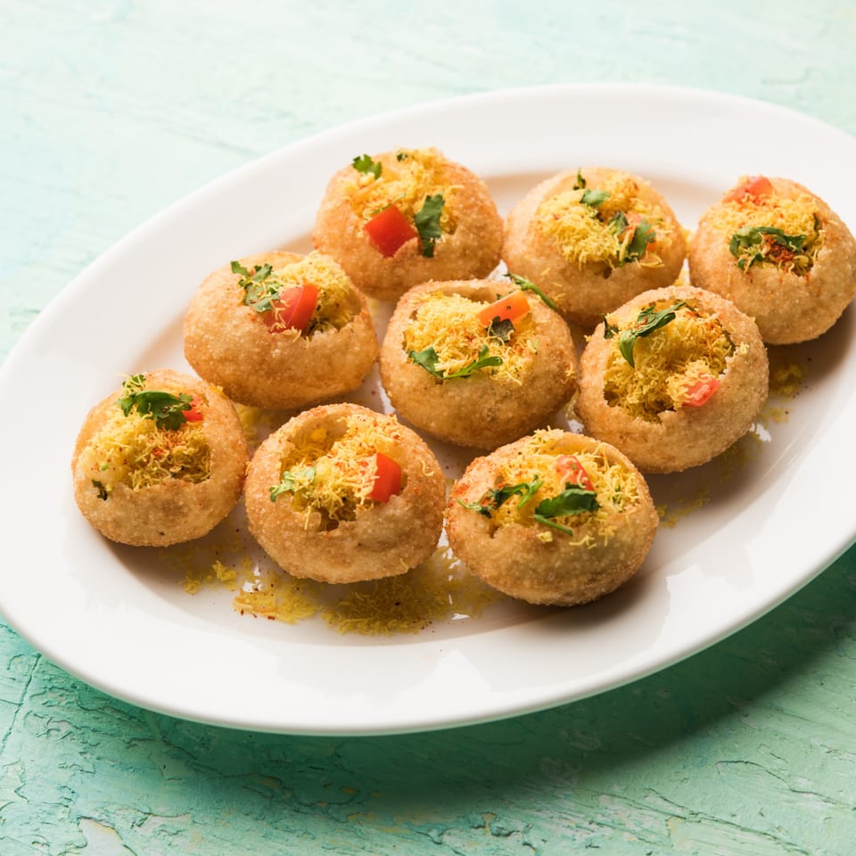A white plate filled with nine Indian pani puri, garnished with sev, cilantro, and diced tomatoes.